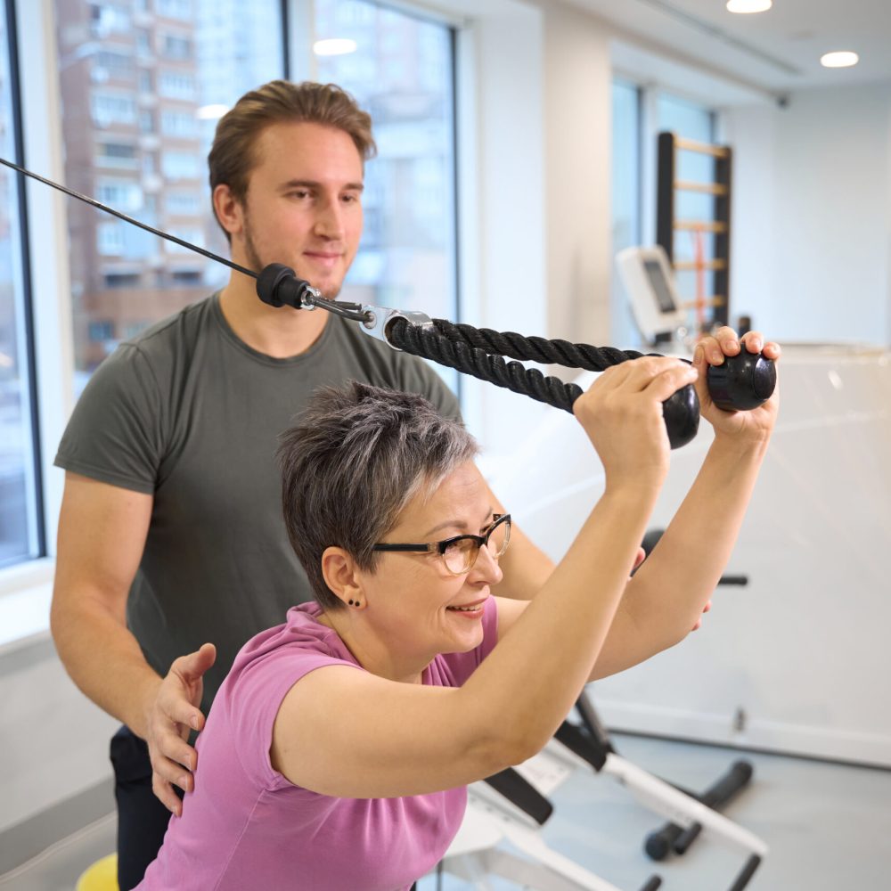 Male trainer teaching smiling adult caucasian woman wearing sportswear doing sport exercise for triceps in gym. Concept of modern healthy lifestyle