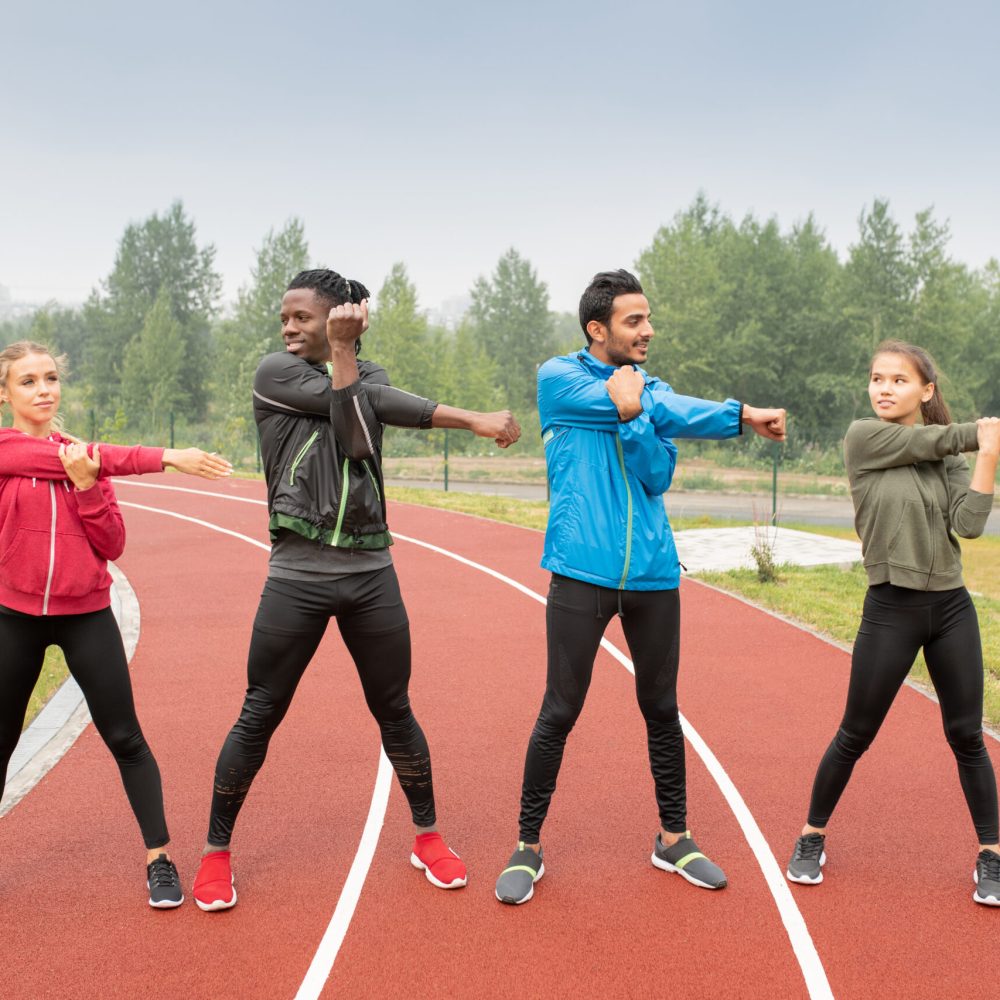 Young men and women in activewear standing on race tracks of outdoor stadium and exercising altogether